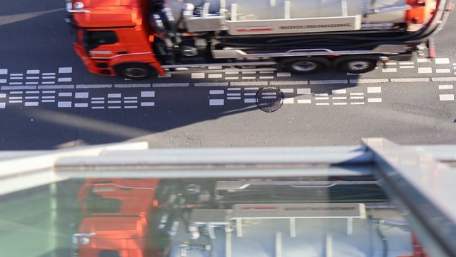 Tankwagen fährt auf einer Straße, gespiegelt in der Glasfassade eines Gebäudes.